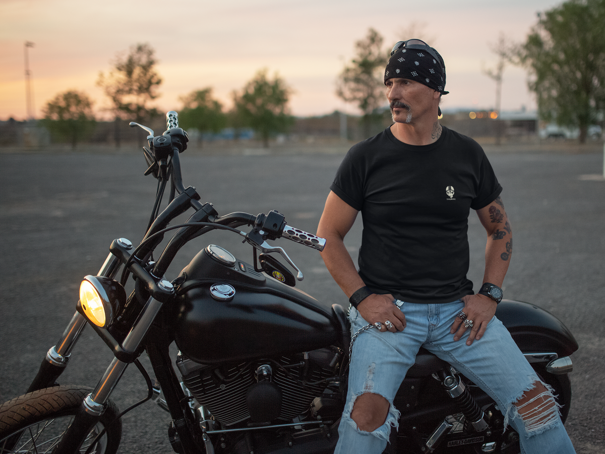Rider in a black StrangeMunkey tee leaning against a motorcycle at sunset
